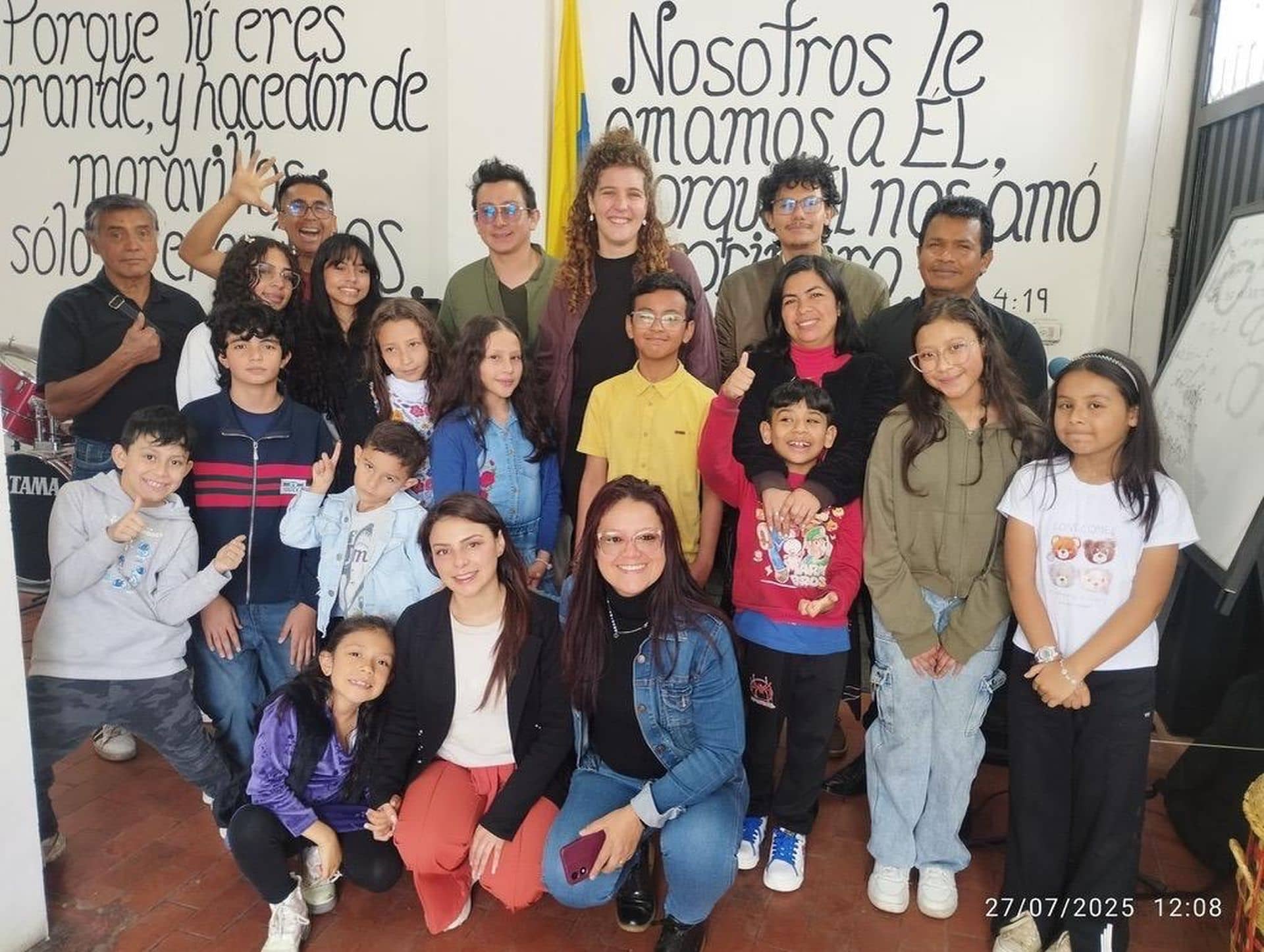A diverse group of people, including children and adults, smiling and posing indoors with wall art and text in the background.