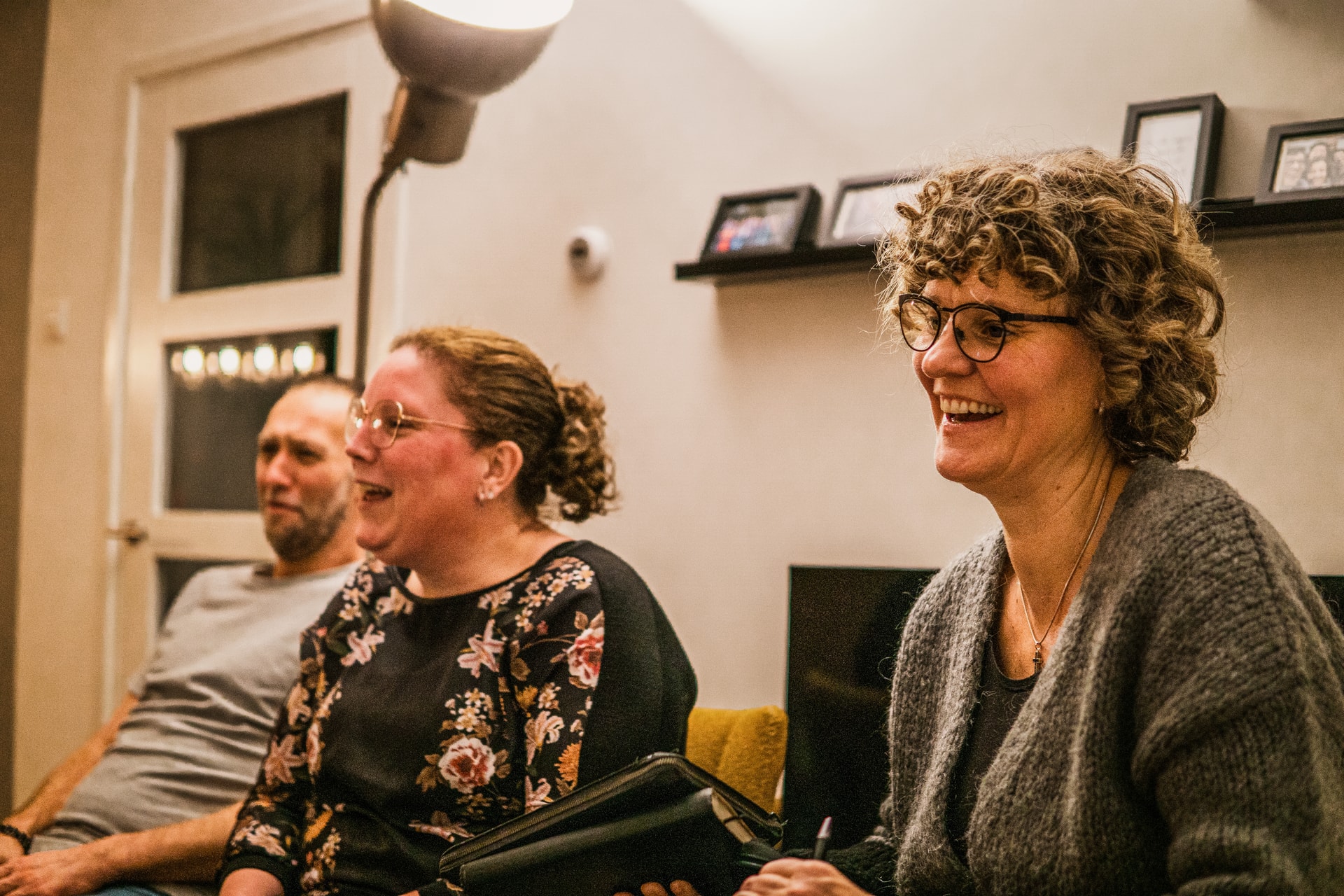 Three people sitting indoors, smiling and laughing. A woman with curly hair holds a notebook. Framed photos are displayed on a shelf behind them.